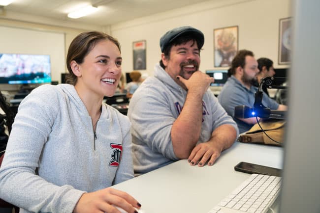 Two students smiling and laughing around an iMac computer in the communication studies class Computer Animation and Voice Acting